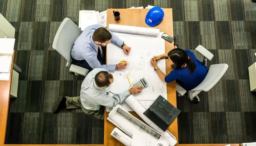 Tres personas analizan planos en una mesa. El significado de este predio se discute con una calculadora y un casco azul.