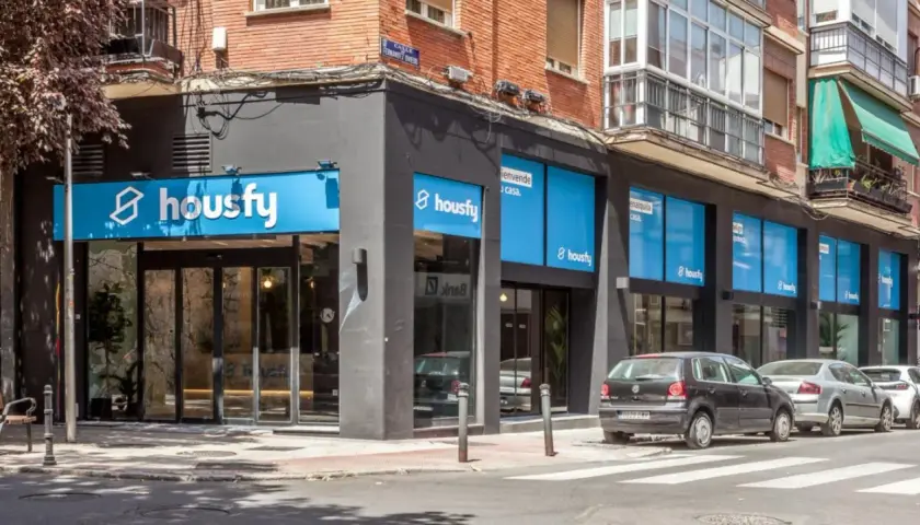 Fachada de oficina de Housfy, con letreros azules y blancos. Las opiniones sobre Housfy son positivas, como se ve en su presencia en la calle.