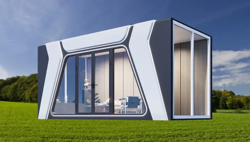 Moderna casa cápsula con grandes ventanales, ubicada en un campo verde bajo un cielo azul.