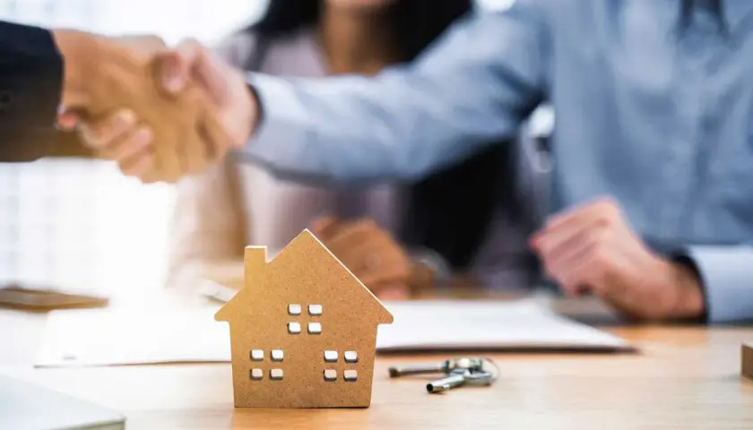 Pareja estrecha manos con agente inmobiliario. Una maqueta de casa y llaves sobre la mesa sugieren que las arras se descuentan de la hipoteca.