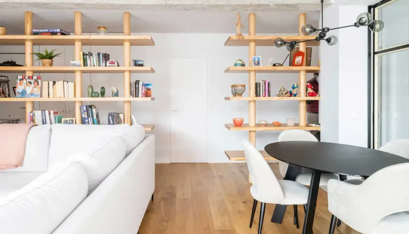 Modern living room with white sofa, wooden shelves filled with books and decor, and a dining area. Un ejemplo de cambio de uso de local a vivienda.