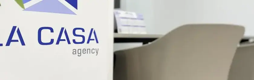 Logo de "LA CASA agency" en un cartel blanco, con sillas de oficina al fondo. Buscando la casa agency opiniones.