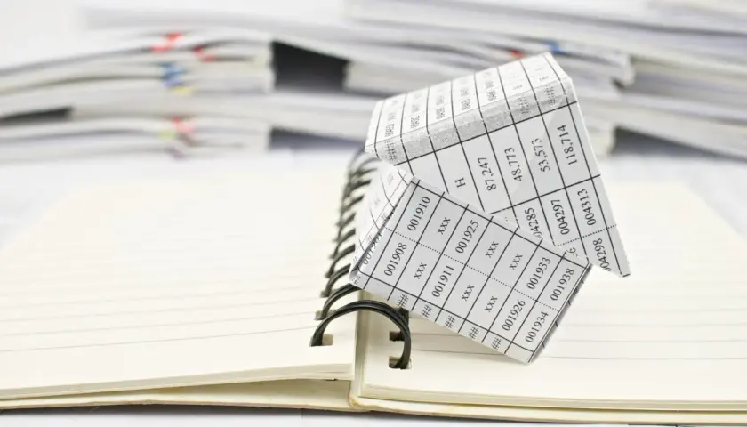 Casa de papel con números, simulando un código registral único, sobre un cuaderno abierto y pilas de documentos.