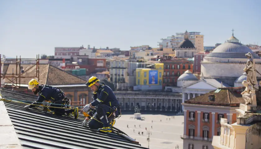 Dos operarios con arnés y casco amarillo trabajan en un tejado, con la ciudad de Nápoles de fondo. Tienen el permiso para cambiar tejado.