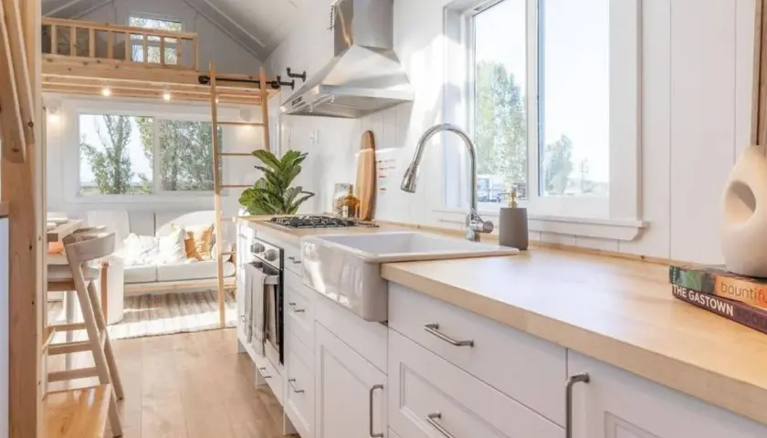 Cocina luminosa y funcional de una tiny house, con encimeras de madera, fregadero blanco y una acogedora sala de estar al fondo.
