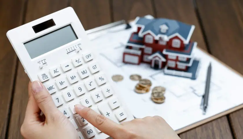 Manos calculando con calculadora blanca, casa modelo, monedas y bolígrafo. Planificando cómo desgravar alquiler.