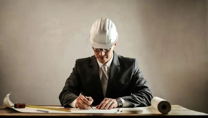 Un aparejador que es, con casco y traje, dibuja planos en su escritorio.