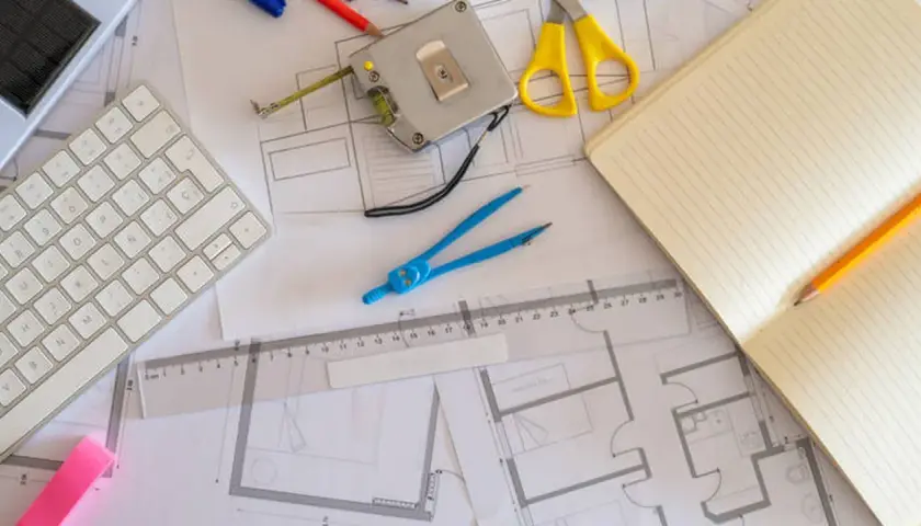 Herramientas de diseño sobre una superficie gráfica: teclado, cinta métrica, tijeras, compás, regla, cuaderno y lápiz.