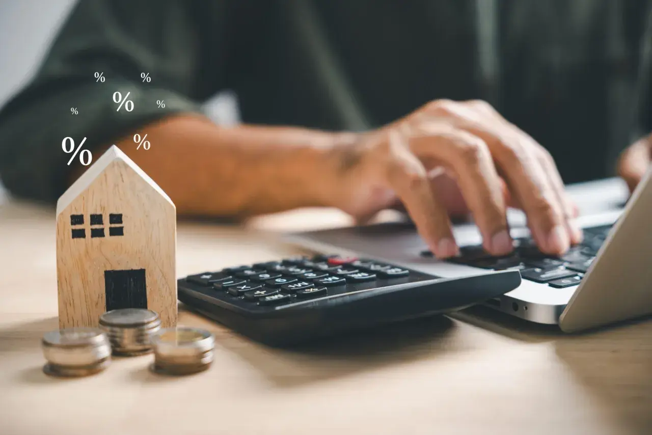 Calculadora, monedas y casa de madera junto a un portátil. Símbolos de porcentaje flotan, sugiriendo el cálculo del precio vivienda.