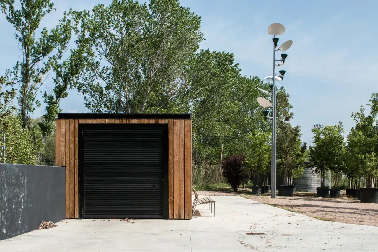 Exterior de una casa de aperos con puerta enrollable negra y revestimiento de madera. Un banco metálico y una farola moderna completan la escena.