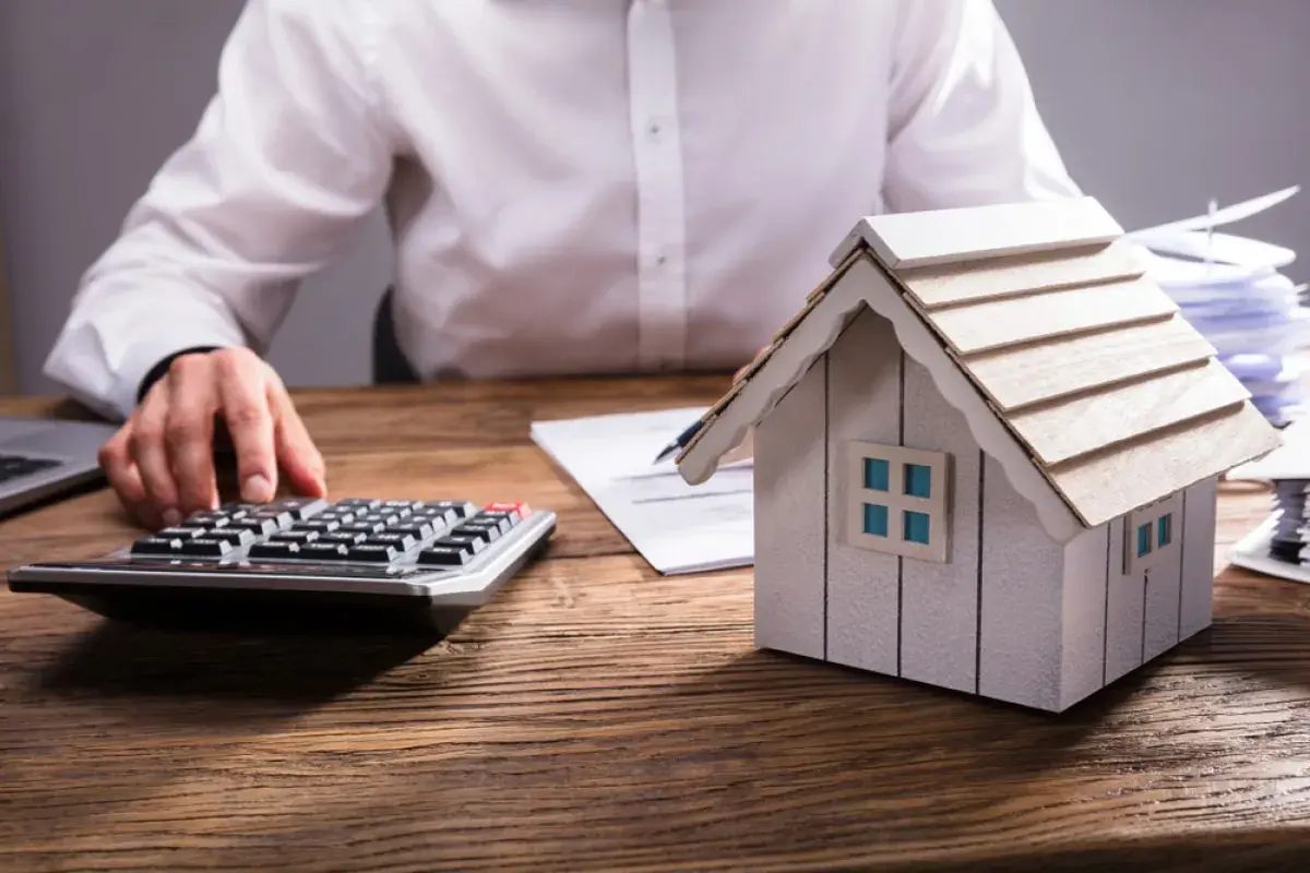 Persona calculando el valor de una propiedad, con una maqueta de casa, calculadora y documentos. Aprende como tasar una vivienda.