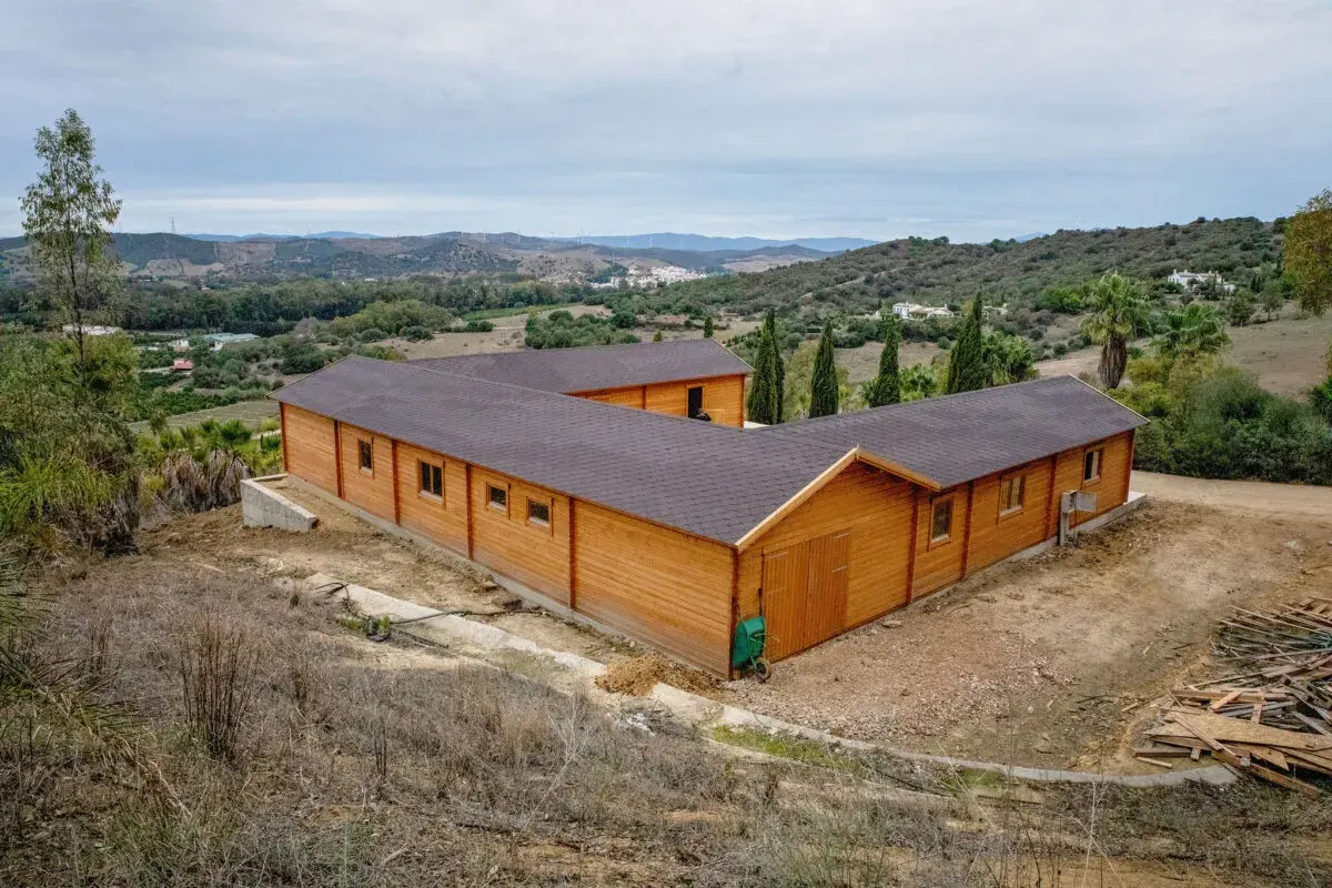 Edificio de madera en forma de L, que es una finca rústica, rodeado de vegetación y colinas.