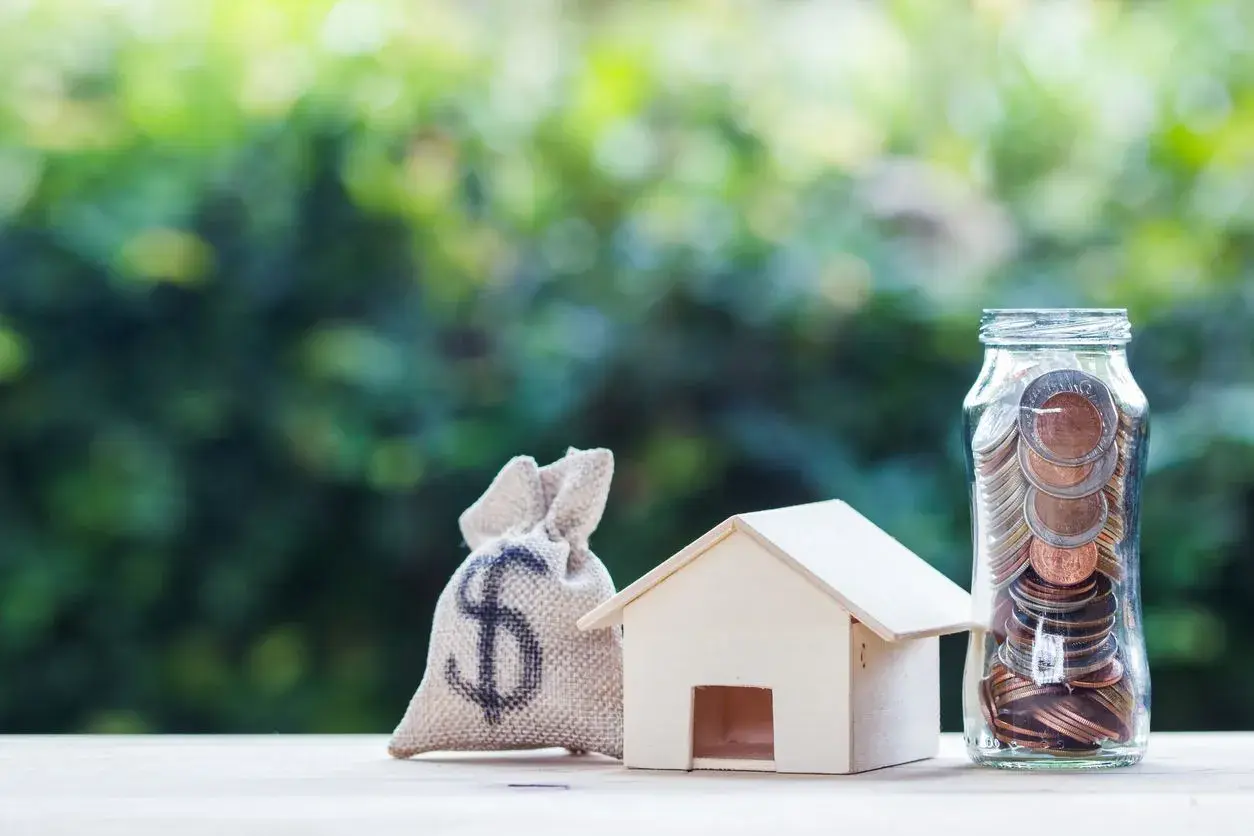 Saco de dinero, casa de madera y frasco lleno de monedas, simbolizando el pleno dominio de tus finanzas para adquirir tu hogar.