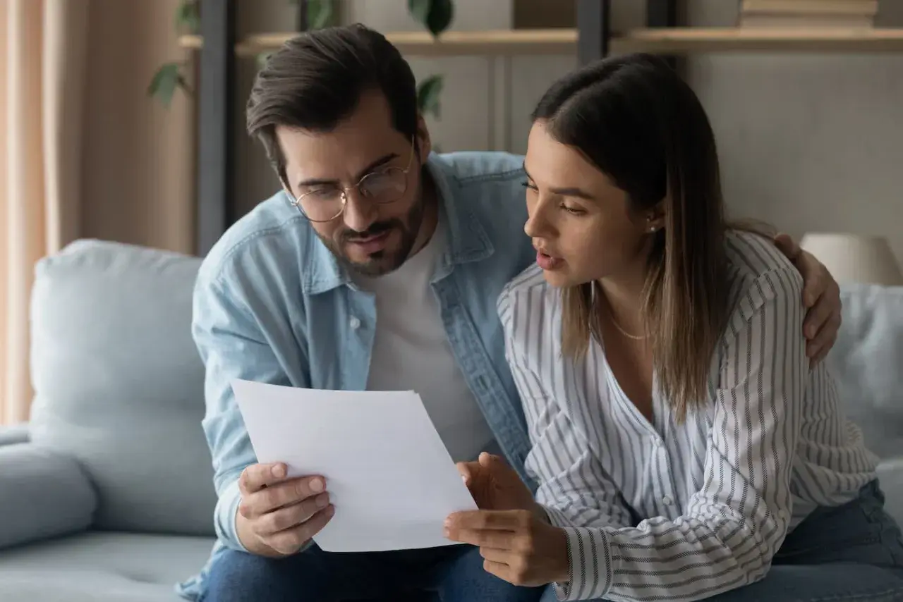 Pareja revisando un documento, el hombre con gafas y camisa vaquera, la mujer con camisa a rayas. Él le pone una mano en el hombro, mostrando apoyo.