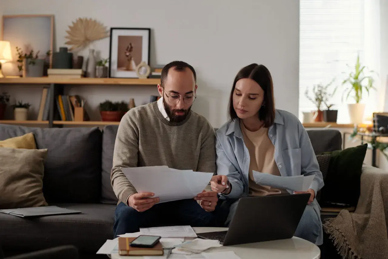 Pareja revisa documentos y laptop, debatiendo qué tasadora tasa más alto para su hipoteca.
