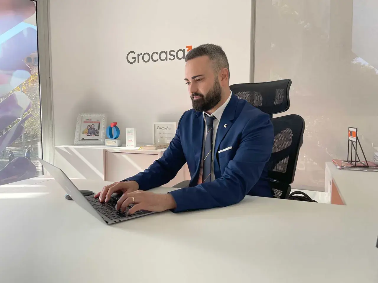 Hombre de negocios trabajando en su portátil en una oficina con el logo de Grocasa. Busca opiniones sobre Grocasa para su próximo proyecto.