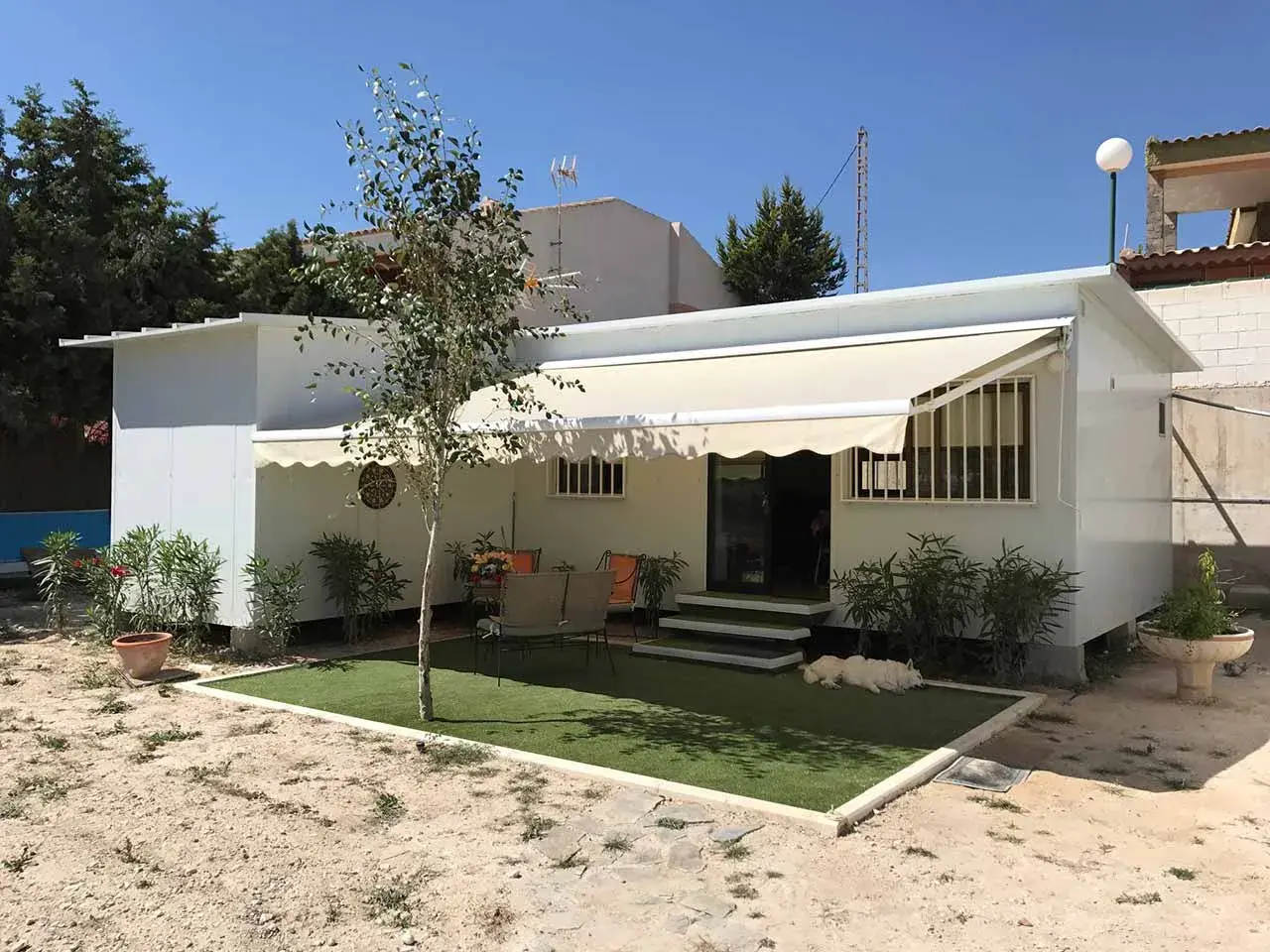 Una moderna casa de aperos blanca con toldo, césped artificial y un perro blanco descansando.