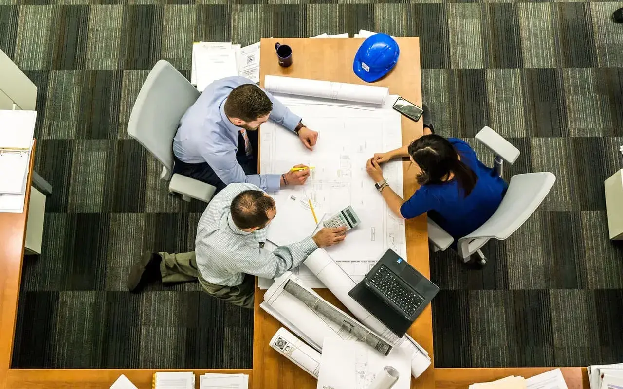 Tres personas analizan planos en una mesa. El significado de este predio se discute con una calculadora y un casco azul.