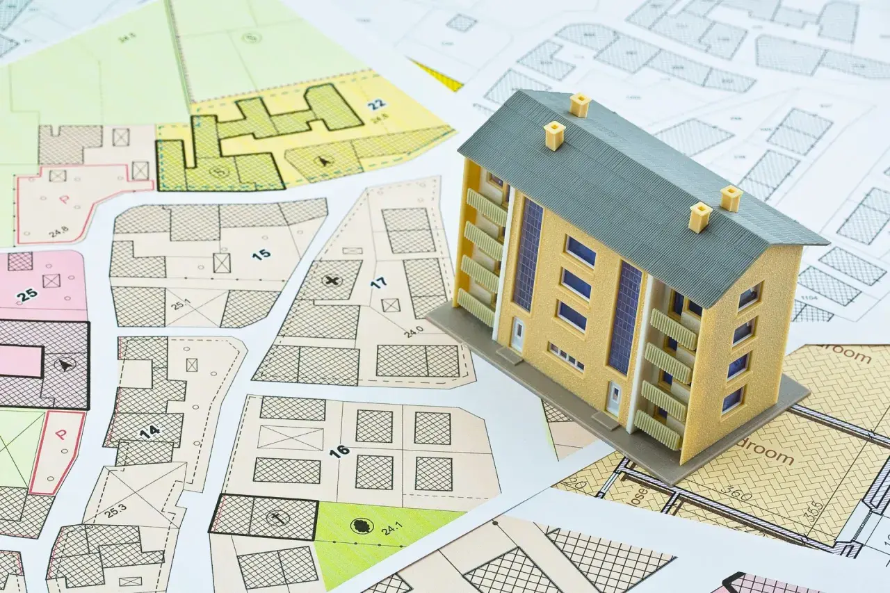 Maqueta de vivienda sobre planos urbanísticos. El modelo representa un edificio de varias plantas, con balcones y ventanas. Los planos detallan parcelas y edificaciones, sugiriendo la planificación para determinar el **valor catastral vivienda**.