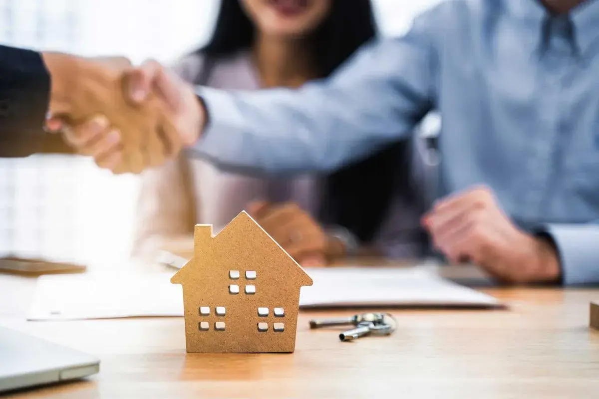 Pareja estrecha manos con agente inmobiliario. Una maqueta de casa y llaves sobre la mesa sugieren que las arras se descuentan de la hipoteca.
