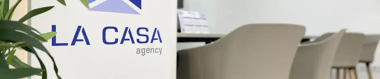 Logo de "LA CASA agency" en un cartel blanco, con sillas de oficina al fondo. Buscando la casa agency opiniones.
