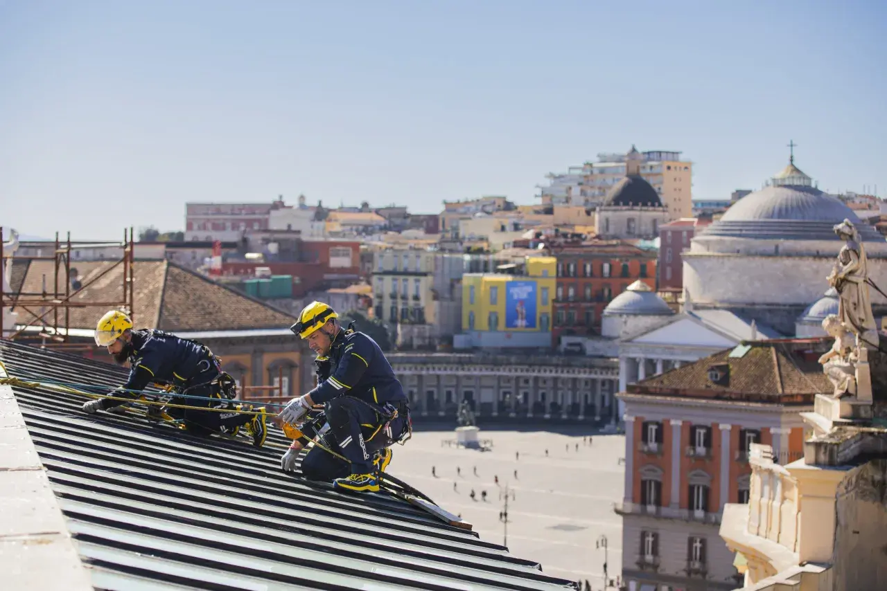 Dos operarios con arnés y casco amarillo trabajan en un tejado, con la ciudad de Nápoles de fondo. Tienen el permiso para cambiar tejado.