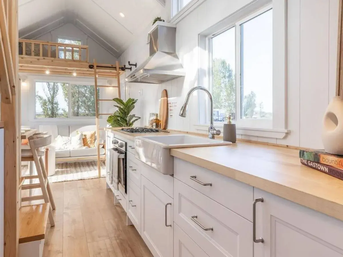 Cocina luminosa y funcional de una tiny house, con encimeras de madera, fregadero blanco y una acogedora sala de estar al fondo.