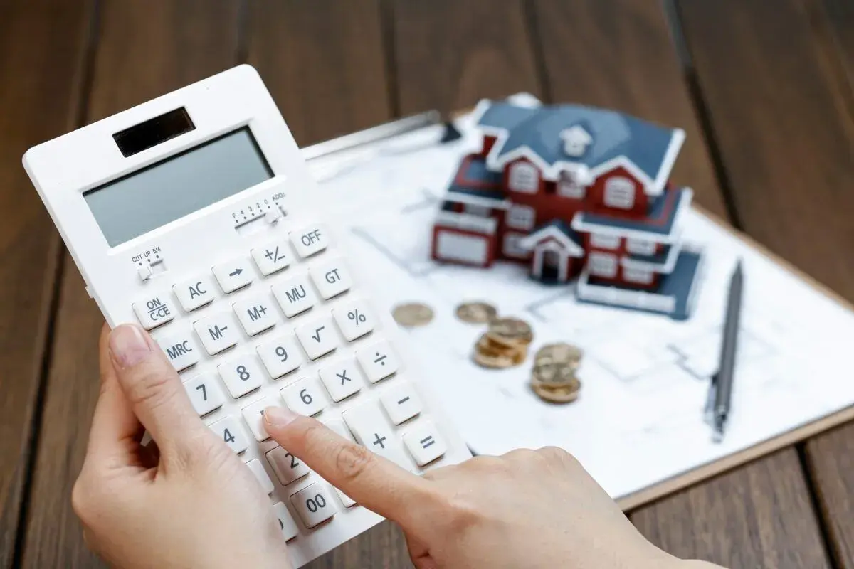 Manos calculando con calculadora blanca, casa modelo, monedas y bolígrafo. Planificando cómo desgravar alquiler.