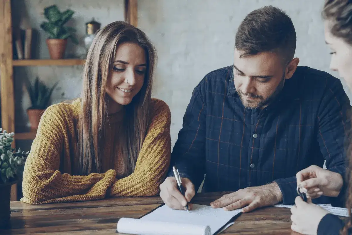 Pareja sonriente firma documentos para una hipoteca sin contrato fijo. Él firma, ella sonríe y le da las llaves.