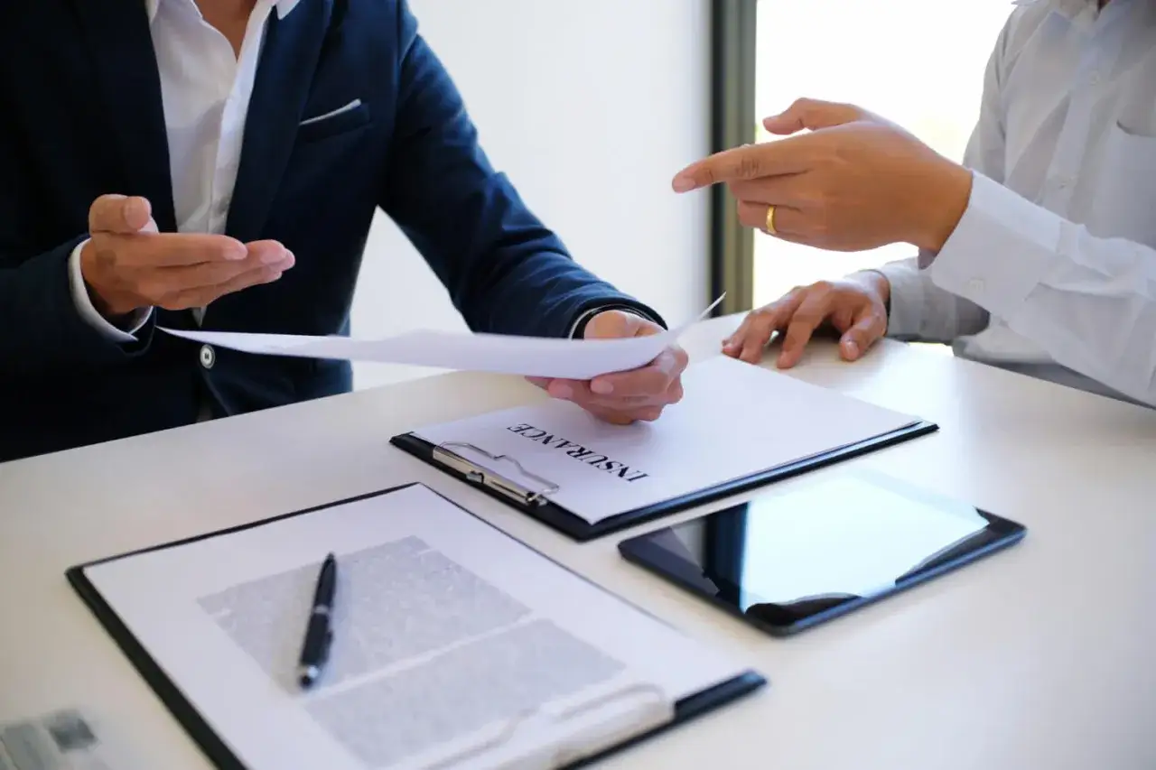 Dos hombres discuten documentos en una mesa. Uno entrega un papel al otro, quien señala. Posiblemente brokers hipotecarios revisando un contrato.