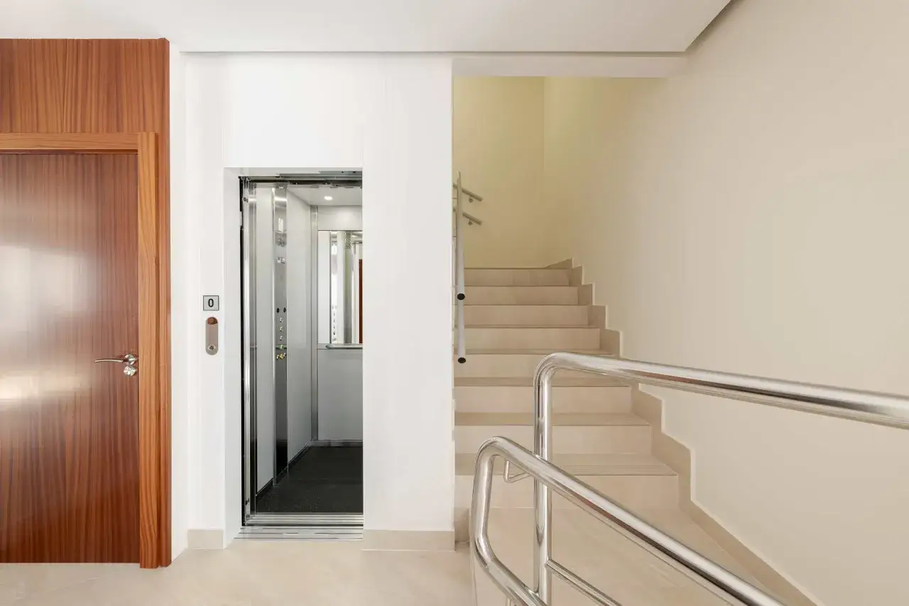 Puerta de madera, ascensor abierto y escaleras con barandilla metálica, elementos comunes de un edificio moderno.