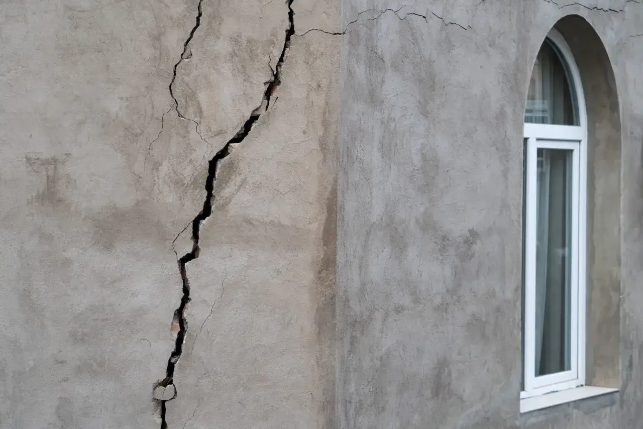 Una grieta profunda atraviesa el muro de estuco, revelando un vicio oculto. A la derecha, una ventana arqueada blanca contrasta con la textura rugosa.