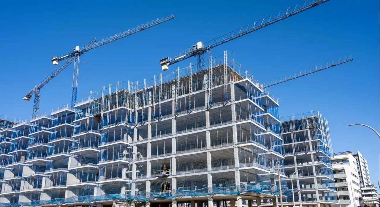 Grúas gigantes supervisan el proceso de construir casa, un edificio de hormigón con múltiples niveles y balcones en construcción.