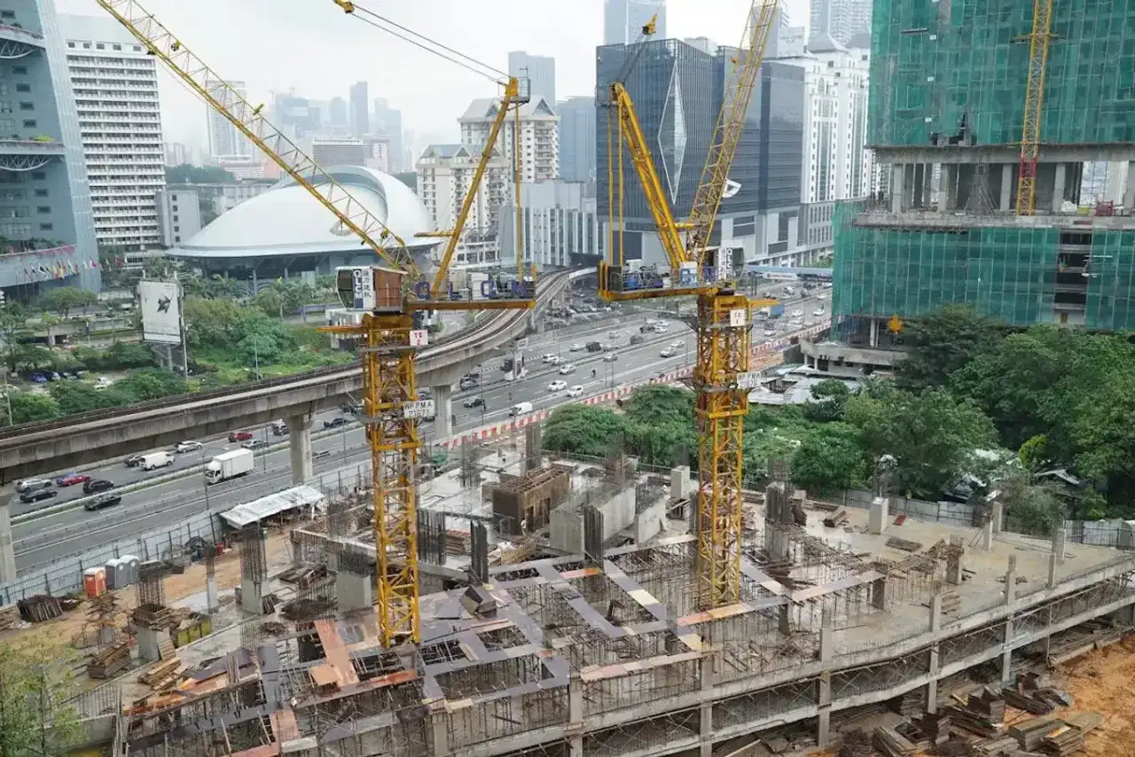 Gr&uacute;as amarillas dominan la escena de construcci&oacute;n de un solar urbano. En el fondo, edificios modernos y una autopista con tr&aacute;fico.