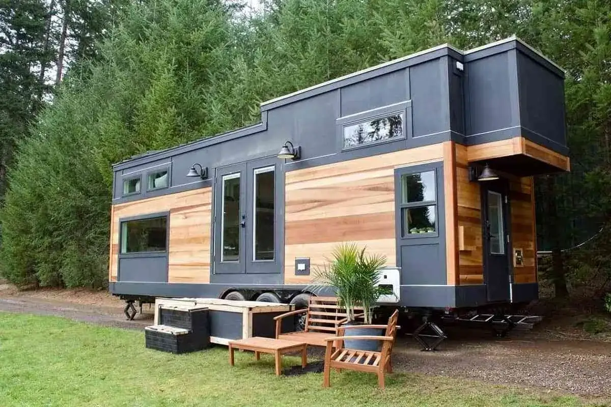 Moderna tiny house con revestimiento de madera y metal, rodeada de árboles. Muebles de exterior invitan a relajarse.