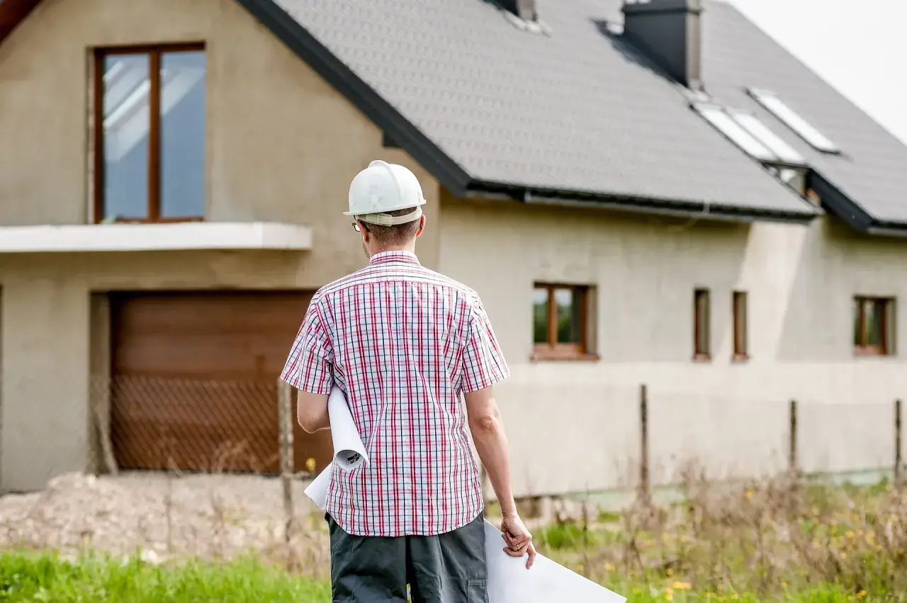 Un aparejador con casco blanco y camisa a cuadros revisa planos frente a una casa en construcción.
