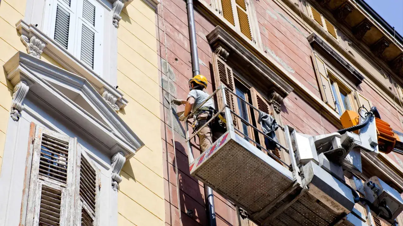 Trabajador en plataforma elevadora realiza mantenimiento de edificios, reparando la fachada con yeso.