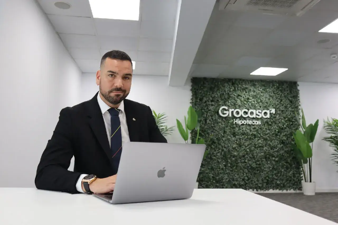 Hombre de traje trabajando en portátil. Detrás, un muro verde con el logo de Grocasa Hipotecas. Buscando grocasa opiniones para tu hipoteca.