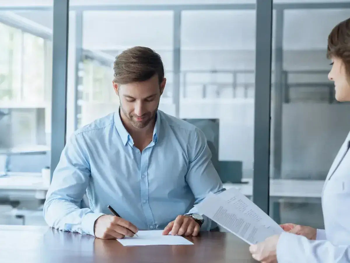 Hombre firma contrato en oficina, mientras mujer revisa documentos. Un ejemplo de servicio en Tecnocasa, agencia inmobiliaria, con rese&ntilde;as positivas.