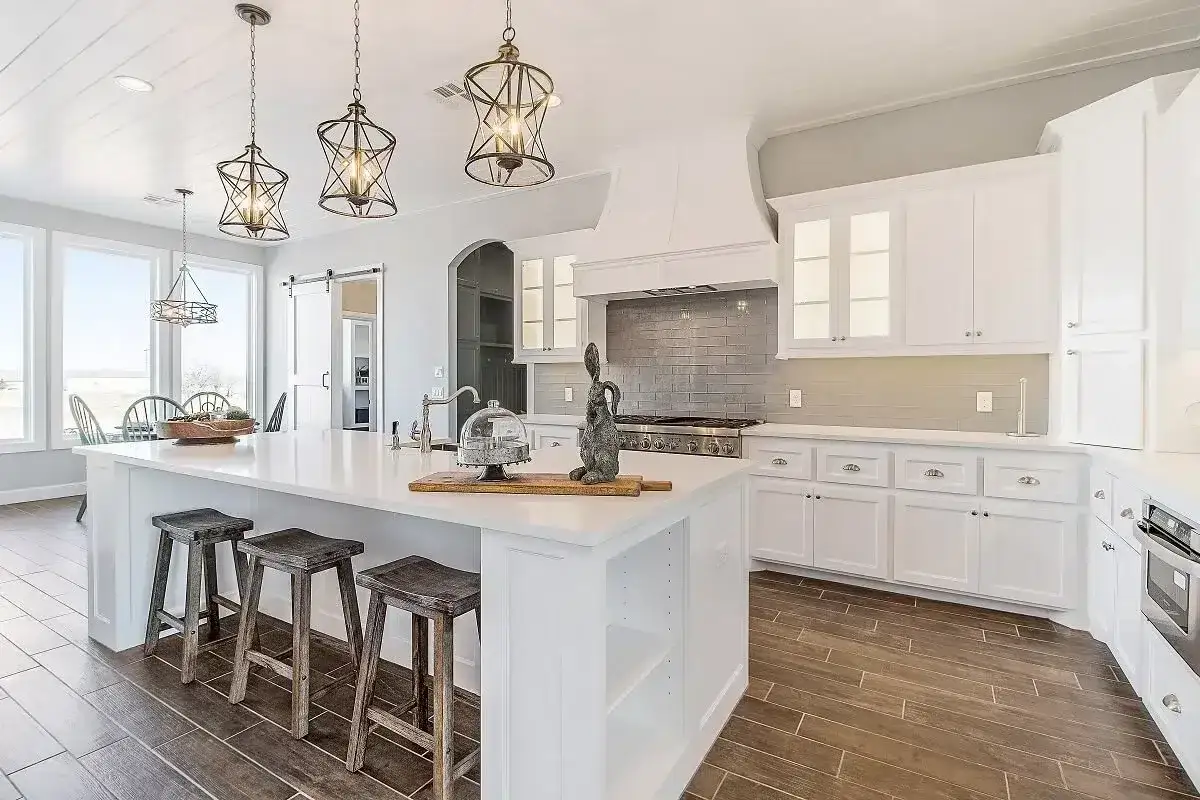 Cocina moderna con isla central, taburetes de madera y lámparas colgantes. Amplia superficie construida con gabinetes blancos y encimeras de cuarzo.