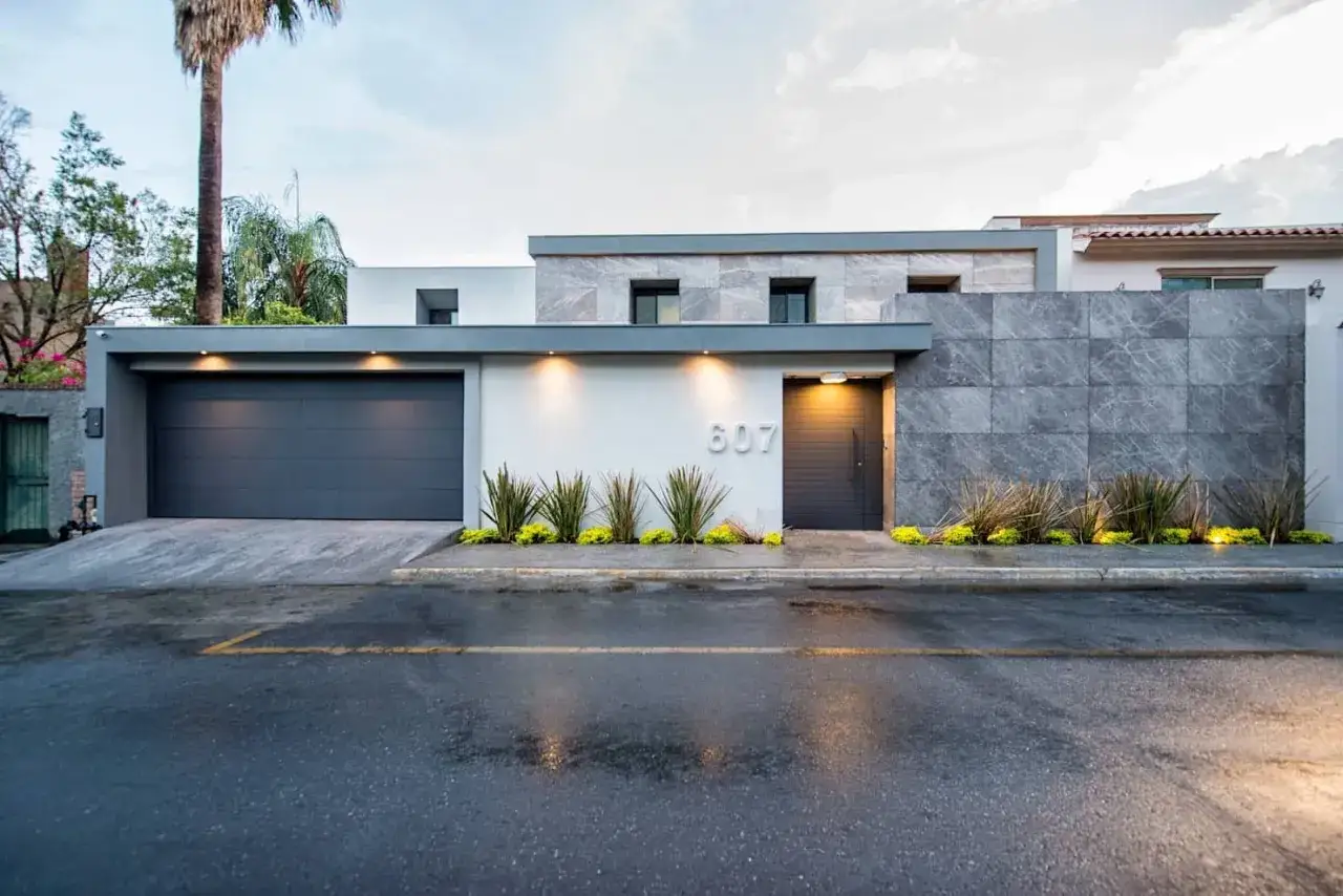 Fachada moderna con portón de garaje gris, puerta de entrada oscura y muros de piedra. Diversos tipos de cerramientos exteriores realzan su diseño.