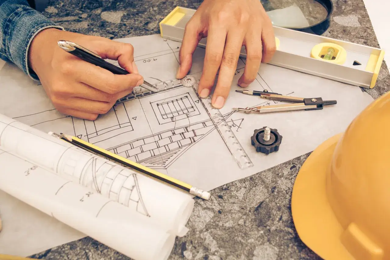 Manos trabajando en el levantamiento de planos, con herramientas de arquitectura y un casco amarillo.