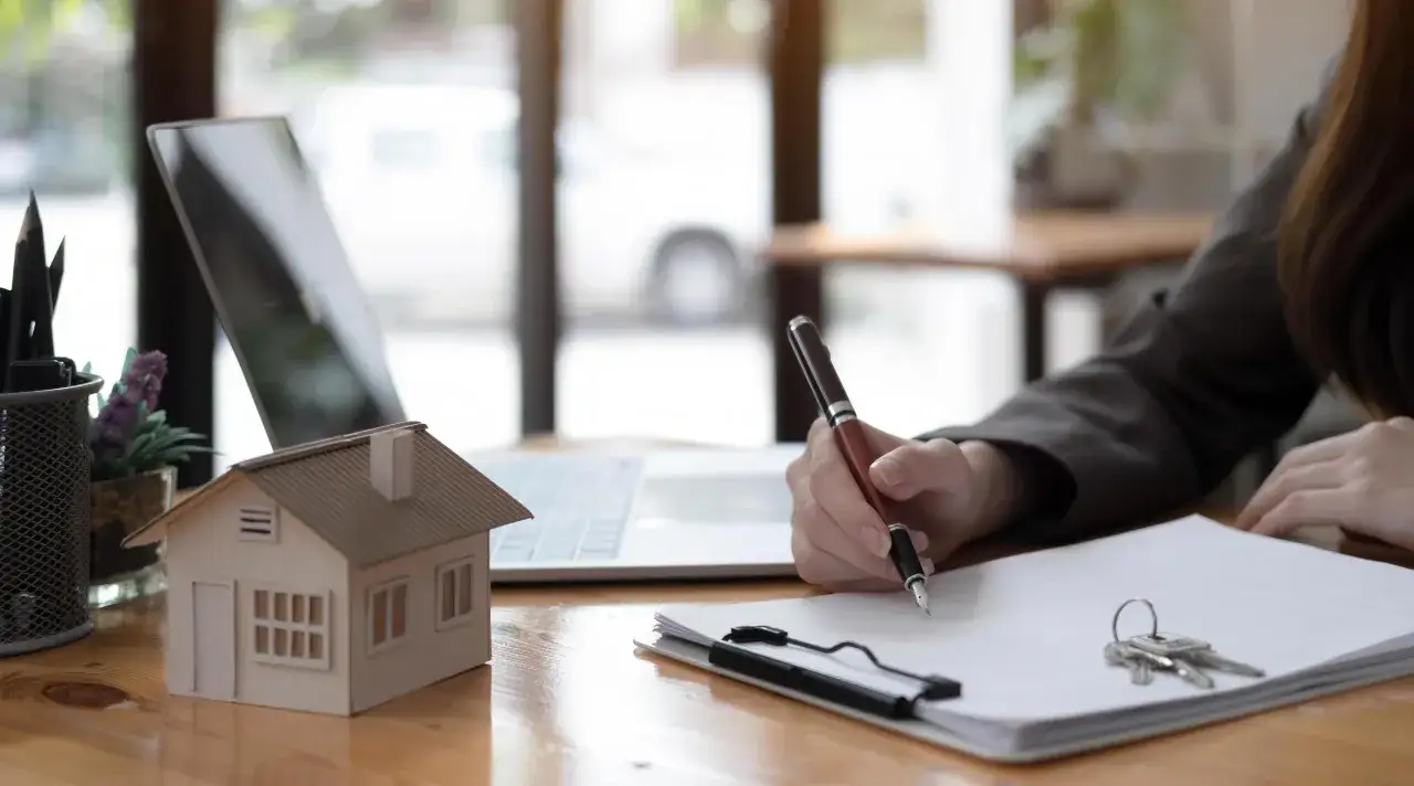 Persona firma contrato de compraventa de un red piso. Laptop, llaves y maqueta de casa sobre escritorio.