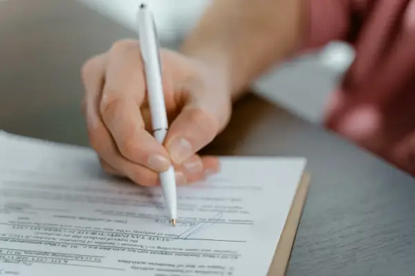 Mano firmando un documento, posiblemente un acuerdo de dación en pago entre particulares, con un bolígrafo blanco sobre papel.
